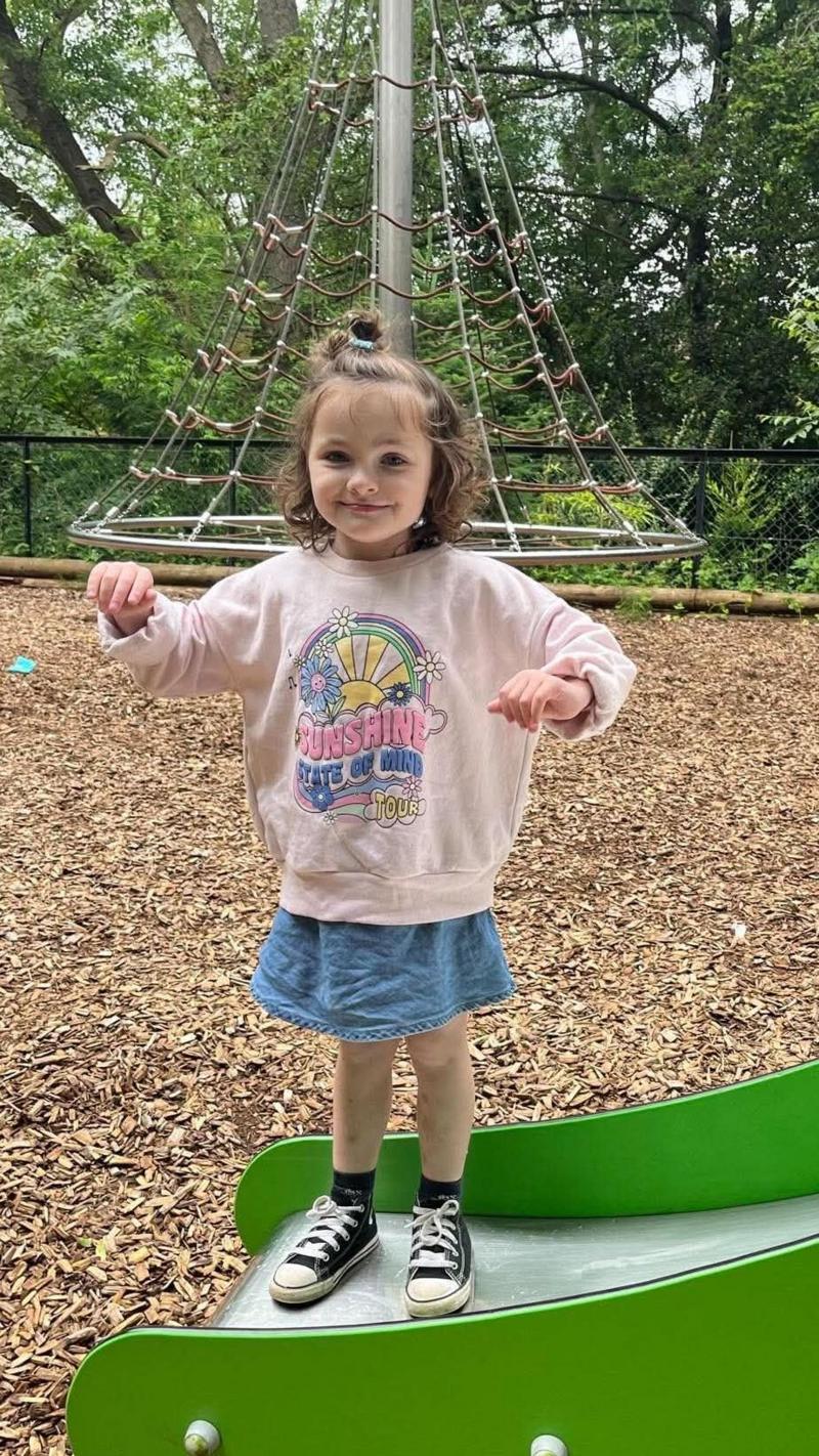 Flora stands on a bright green curved playground balance structure in a wooded play area covered with wood chips.
