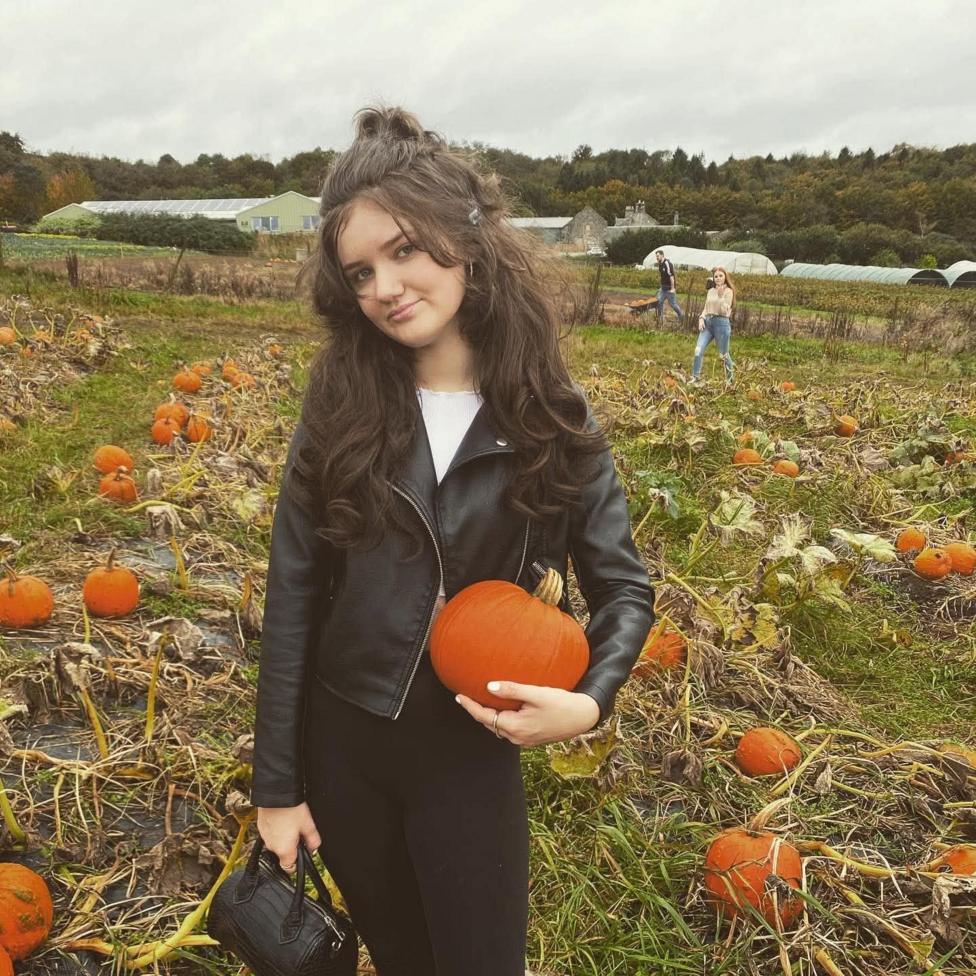 Sophie standing in a pumpkin patch holding a medium-sized orange pumpkin. The person is wearing a black leather jacket and black trousers and carrying a small black handbag.