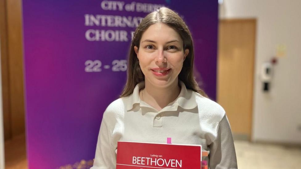 Anna Handler is wearing a white shirt and has long brown hair. She is holding a red book of sheet music, the name of Ludwig Van Beethoven is written on its cover. She is standing in front of a banner promoting the City of Derry Choir Festival