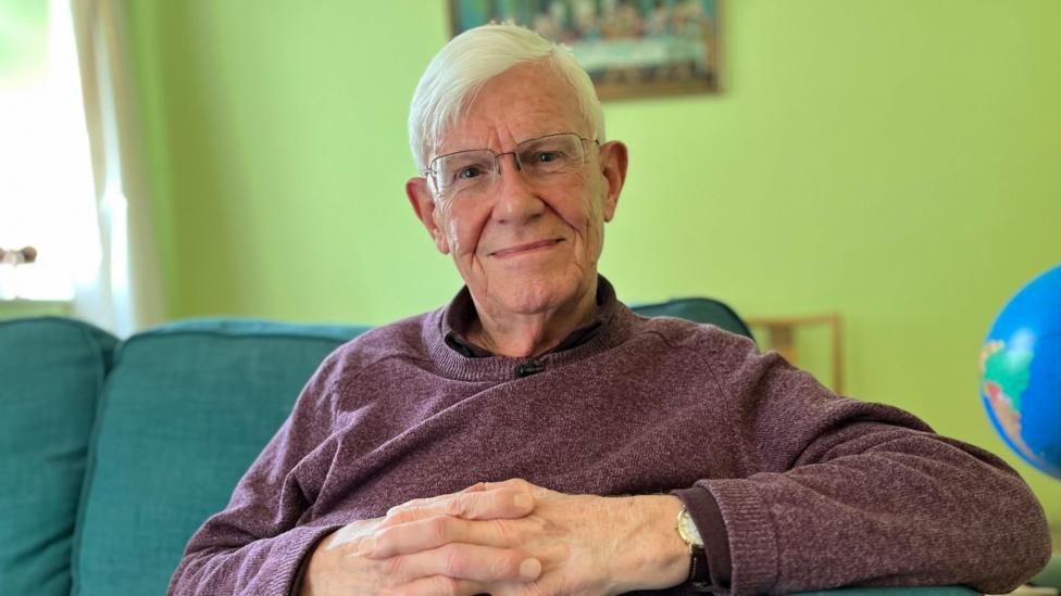 Ken is wearing a purple jumper and glasses. He is looking directly at the camera.