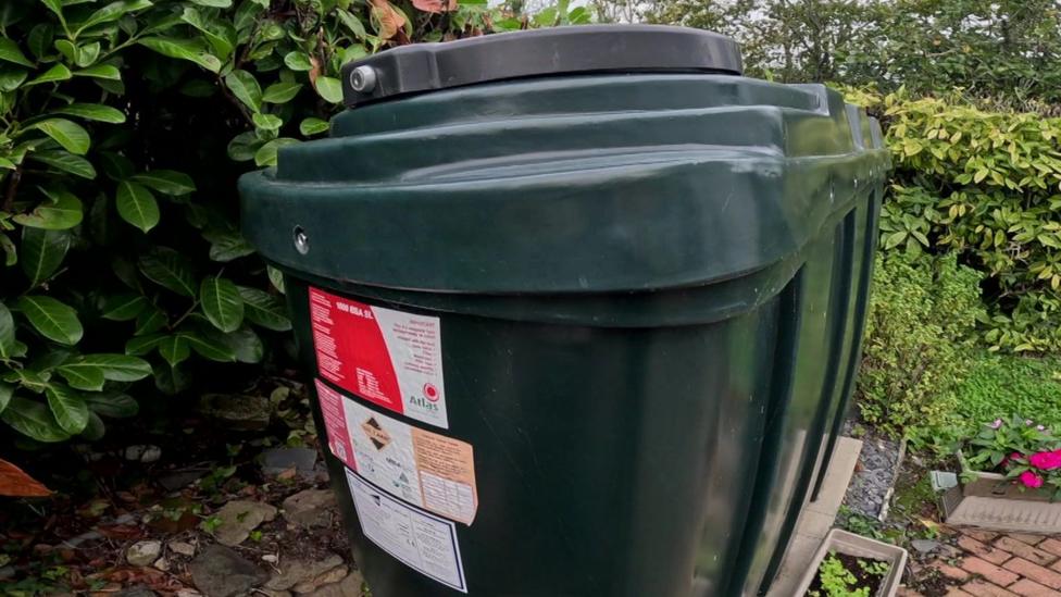This appears to be a large domestic oil storage tank in a garden. The dark green plastic tank has a lockable lid on top and several warning and information labels on the front, which are typical of tanks used to store heating oil for a home.