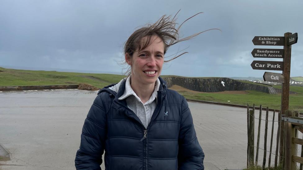 A woman wearing a coat stands with exposed grassy cliffs behind her. Her hair is blowing in the wind.