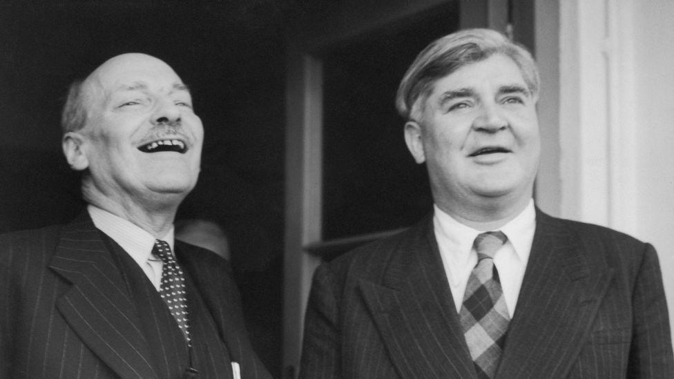 Clement Attlee (left) and Aneurin Bevan (right). Clement throws his head back laughing, while Bevan looks straight ahead, smiling. It is a black and white photo of the two men wearing pinstripe suits, shirts and ties, and with a door open in the background.