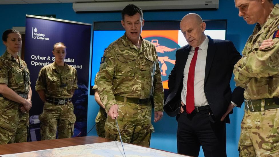 Defence Secretary John Healey is given a briefing as he meets military personnel during a visit to the Permanent Joint Headquarters in Northwood, London. He is wearing a suit and stands alongside military personnel wearing camouflage uniform, with one pointing at a map.