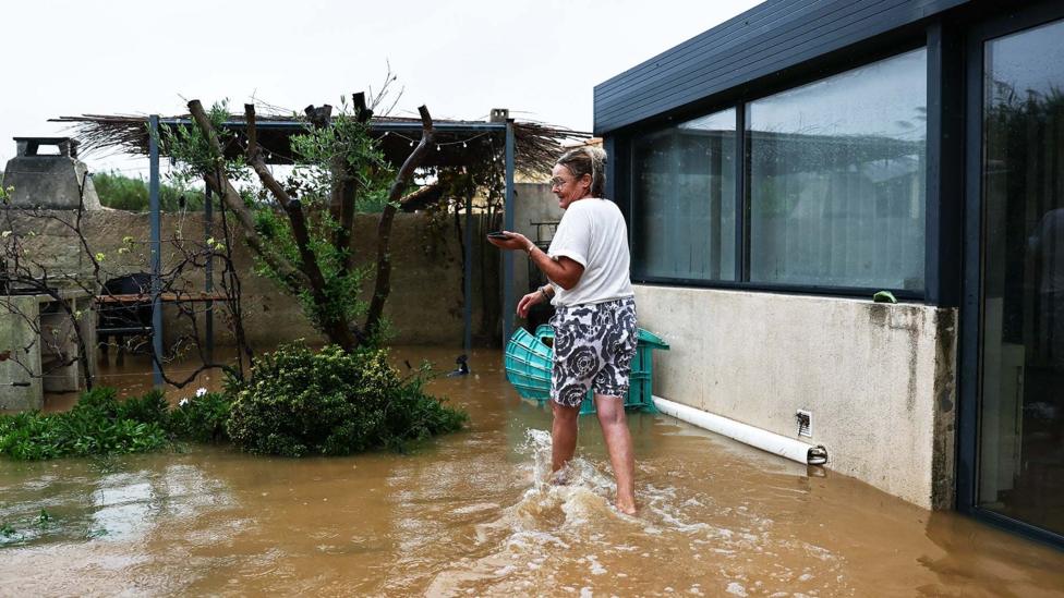 A woman wearing shorts and a white t-shirt wades through brown water in front of her house