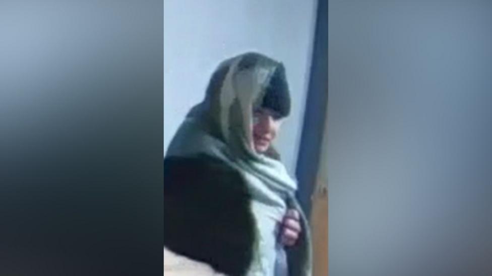 A grainy still from CCTV showing Owens in a corridor in Carla's flat block. He has a green blanket over his head and is wearing a black beanie hat, but his face is exposed and pointing towards the camera.