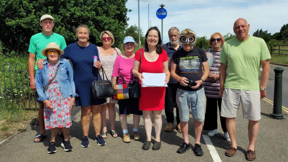A group of 10 people stand looking into the camera smiling. They are all wearing different coloured clothing, including green, red, blue and black. The image was taken on a warm day, the sun is shining on them. The group of people are in shorts and wearing hats. In the middle is Alexandra Kemp, Norfolk County Councillor dressed in a red dress holding up a piece of paper. A man who is standing next to her has an owl mask on his face.