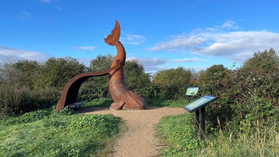 A large bronze statue of a whale's tail positioned amongst greenery.