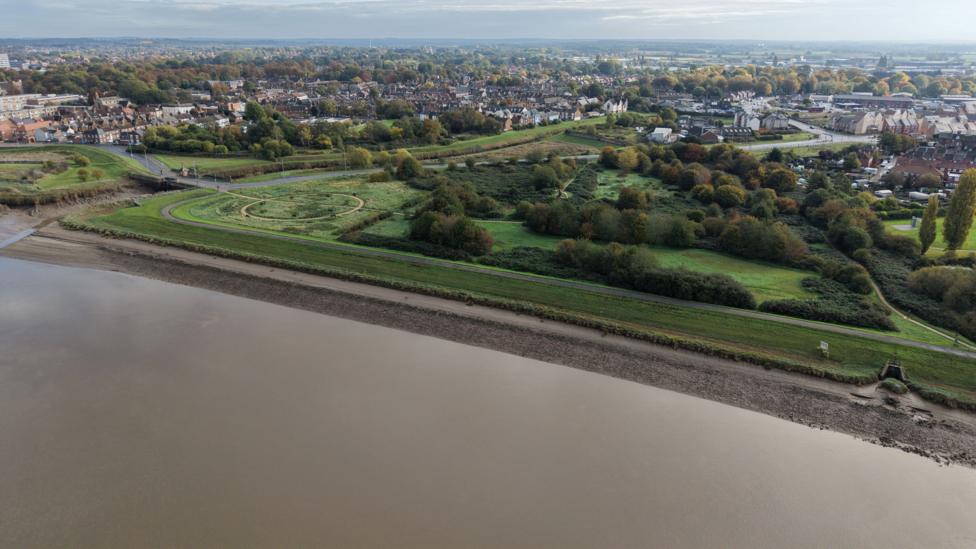 An aerial view of a large green space with a town beyond it. The river is in the foreground.