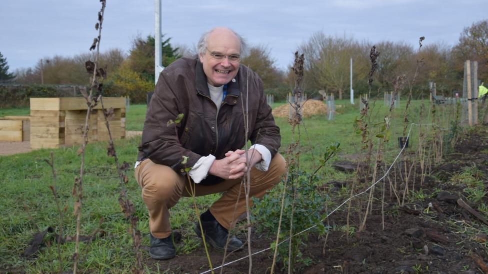 A man in brown jacket and trousers is crouched down on grass, smiling at the camera.