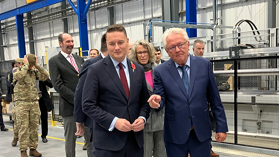 Wes Streeting and another man talk together on a factory floor. There are others in the background, including members of the armed services in uniform