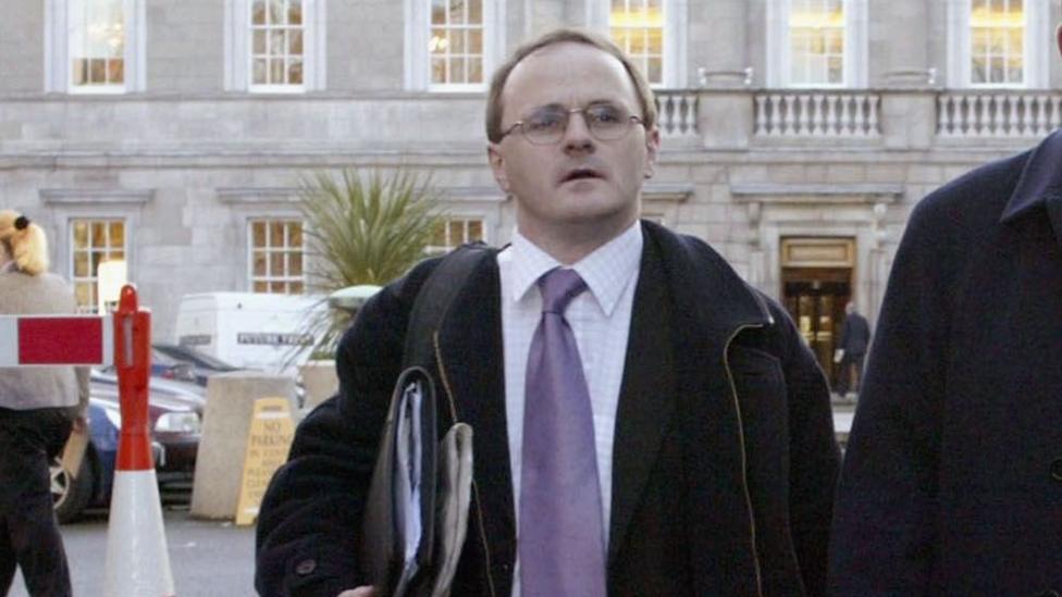 Barry McElduff, pictured outside wearing a black coat, black suit jacket, white shirt and a purple tie. He has short brown hair and wears glasses.