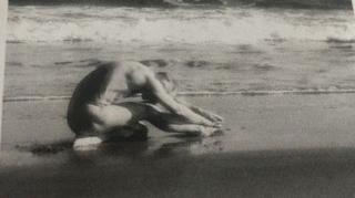 A man sits on a sandy beach, he sits on one leg while the other is stretched out. He is naked and bent over the stretched leg with his arms outstretched. He sits close to the sea. The photo is black and white and grainy.
