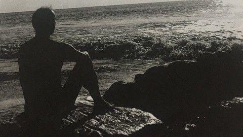 In another black and white photo, Brother Titus is sitting on a rock looking out to sea. He is naked. His knees are bent and he rests his arms on them.