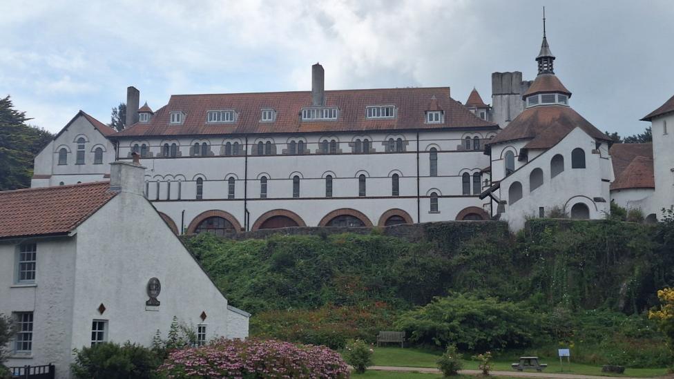 Caldey Abbey is a large white building, which has towers and many windows. The rooves, some of which are conical, are red. There are many windows and it stands on a slight hill overlooking a green and smaller cottages.
