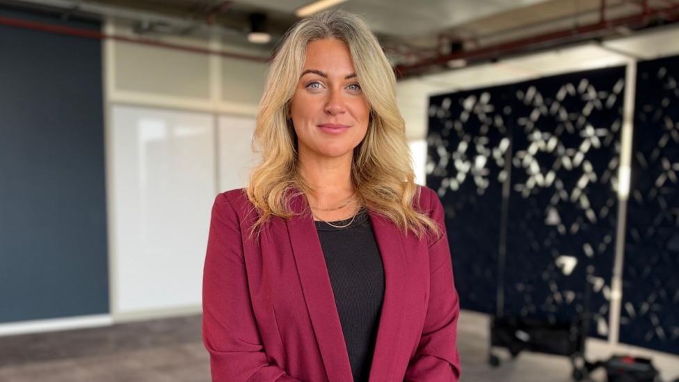 Kayla Poulton, a woman with long blonde hair and blue eyes, smiles in a large room with black patterned screens in the background.