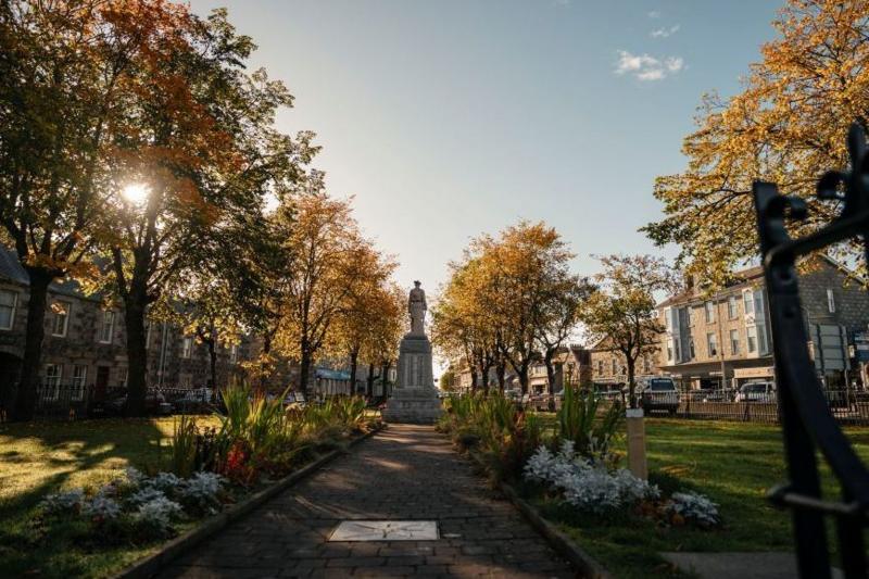 Inverurie town centre, a statue is in the middle of the shot, sunlight is coming through trees, and buildings are on either side.