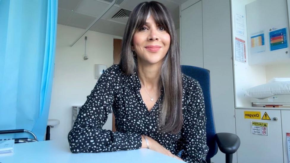 A woman with long brown hair and a fringe smiling, she is sat down on a blue chair and is wearing a black blouse with white polka dots, the background is doctor's clinic.
