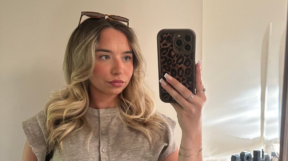 A woman with long blonde hair, she is taking a picture in a mirror with her phone visible in her hand. She is wearing sunglasses on top of her head and a short sleeve beige, button-up top.