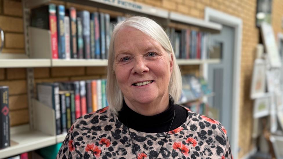 Women with short white hair, standing in front of a bookshelf wearing a black and grey top with red flowers on it.