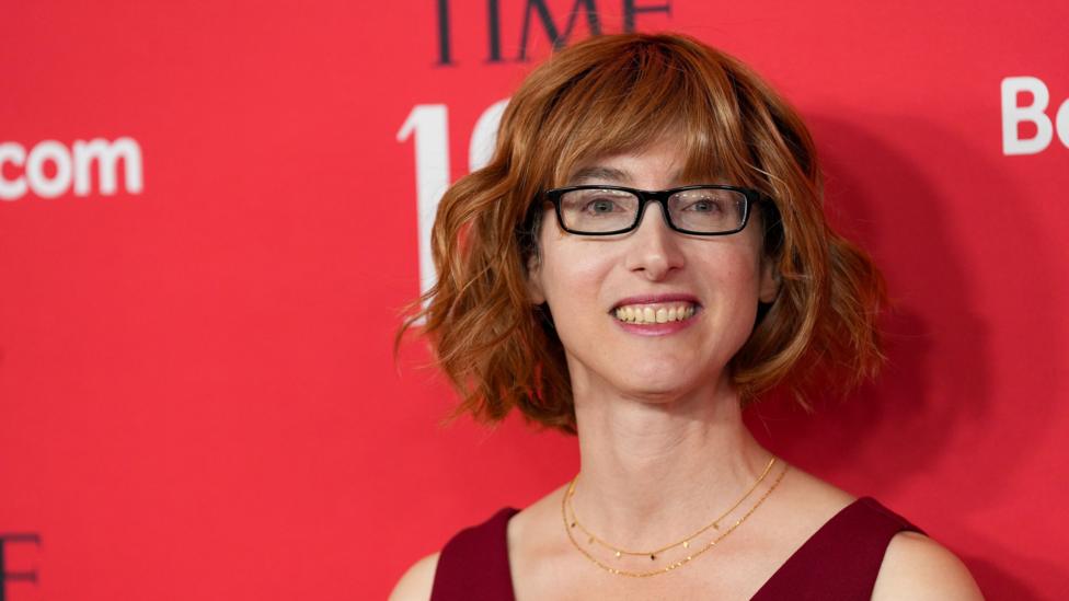 Freida McFadden standing in front of red, black and white background with glasses on