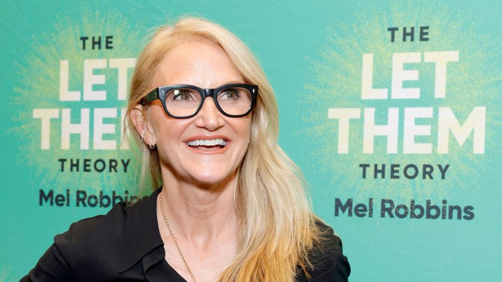 Mel Robbins with long blonde hair wearing a black shirt and glasses. She stands in front of a green background which says 'The Let Them Theory Mel Robbins.'