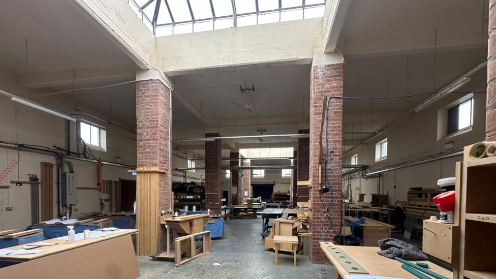 A long, spacious room with high ceilings, red brick columns and two skylights. The room has many wooden workshop tables and shelves.