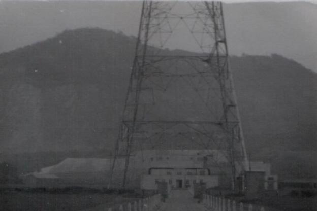 A grainy, black and white photograph of the base of a large radio tower. In the background there is the outline of a large hill that emphasises the size of the tower.