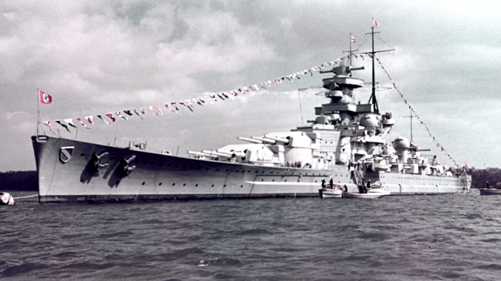 A black and white image of a large warship on the ocean. A garland of flags is strung across the ship, and a red Nazi flag bearing a swastika is on a pole at its helm.