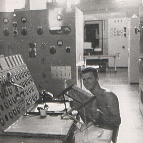 A smiling man sits at a desk holding up a small stool. In front of and behind him is a range of radio equipment.
