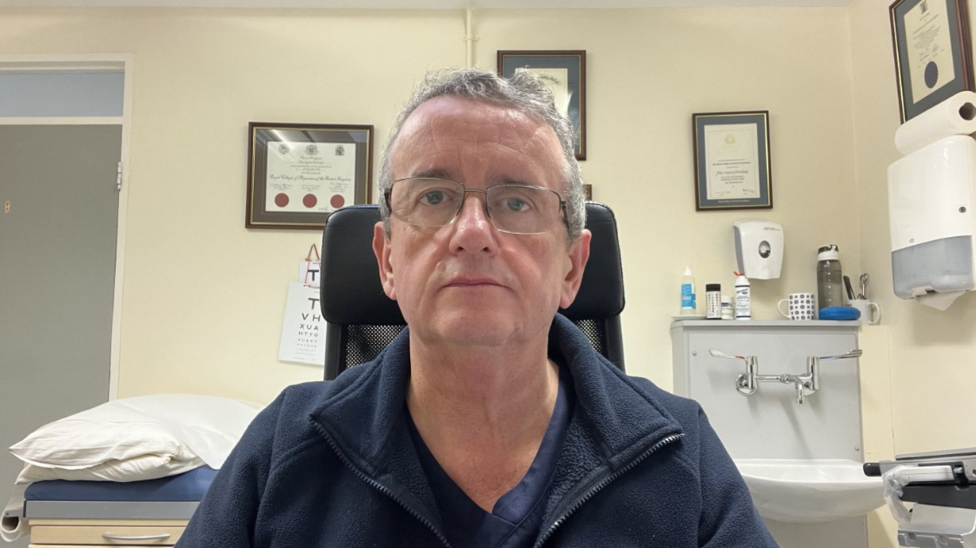 A man with grey hair is facing a camera with glasses, wearing a navy jacket and t-shirt. Behind him is a sink with multiple bottles and a hand sanitiser dispenser sitting on top. The wall features multiple certificates hanging on the wall. To the right of the sink features a hand towel dispenser