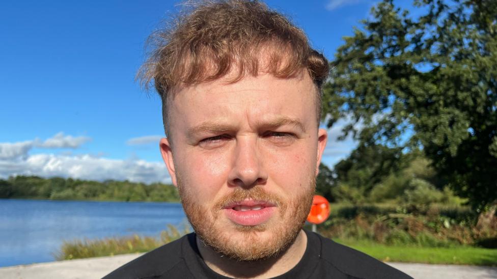 A man with short brown hair and facial hair. He is wearing a black t-shirt and is standing in front of a body of water and some green trees.