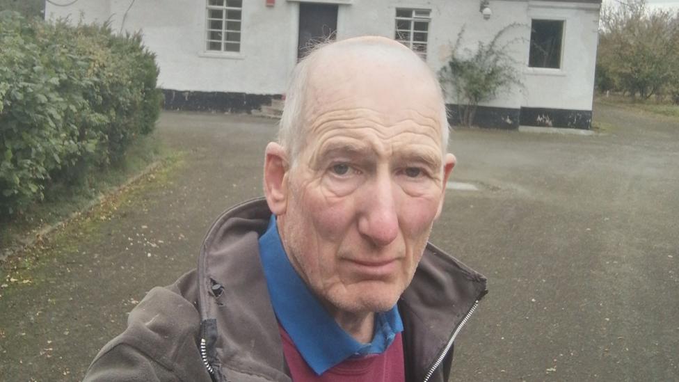 A selfie of Robert Pearce outside a small white building. Robert has short grey hair and is wearing a brown jacket.