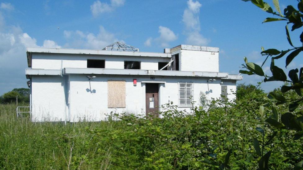 A small, white building with both empty and boarded-up windows, surrounded by overgrown grass and shrubbery.