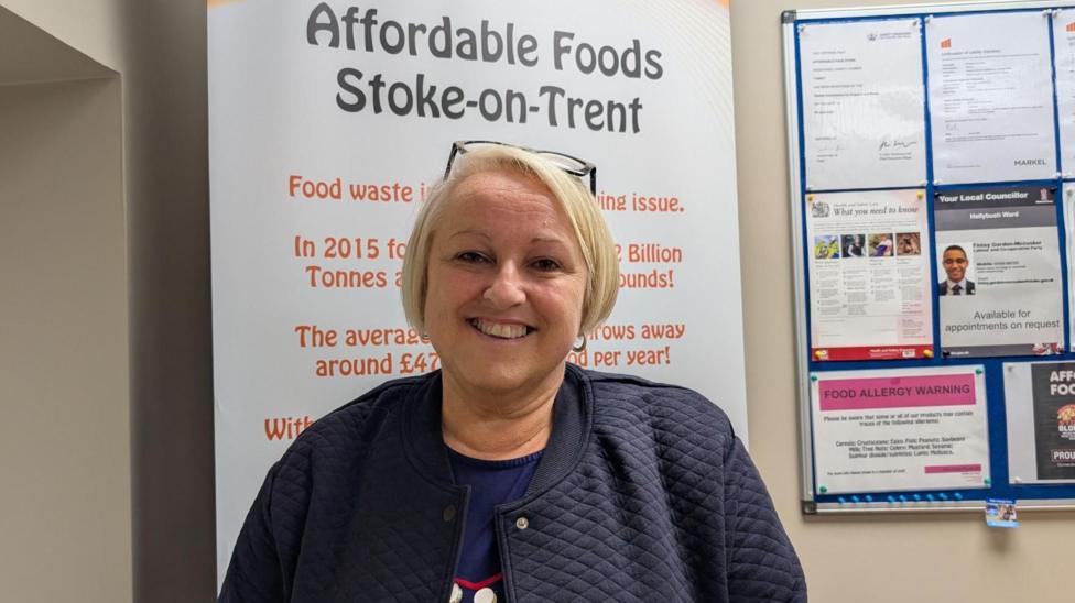 A person wearing a dark quilted jacket stands indoors in front of a display banner reading "Affordable Foods Stoke-on-Trent". A noticeboard containing posters, notices and certificates is visible on the wall behind them.