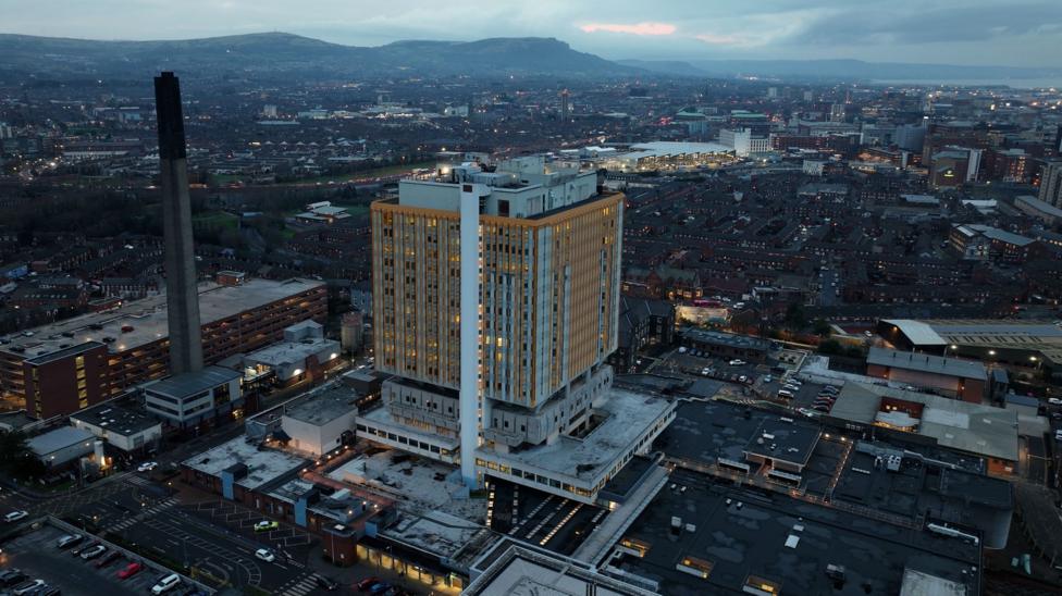 An aerial view of Belfast City Hospital against the city skyline at dusk. The square multi-storey tower block features bright yellow cladding on its exterior. There are hills in the distance.