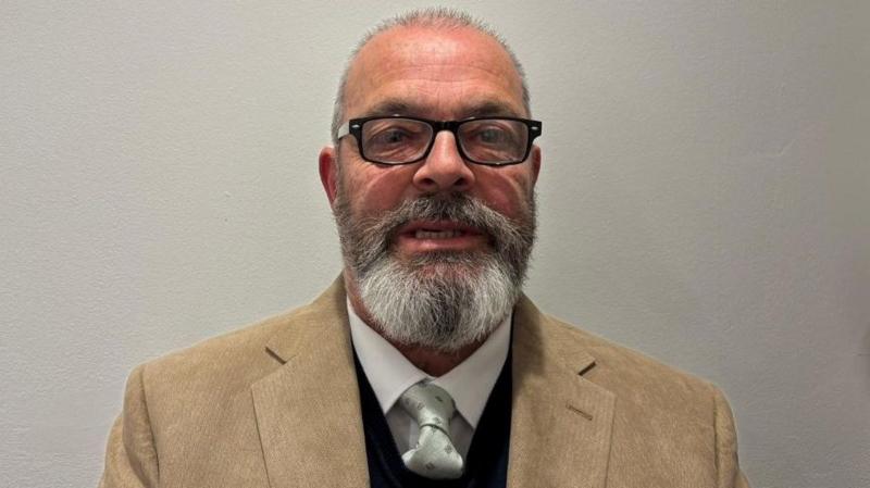 The image shows a man standing against a plain, light-coloured wall. He is facing towards the camera and is wearing black rectangular glasses and has short hair, a full greying beard and moustache. His clothing consists of a tan blazer worn over a dark sweater, with a white shirt underneath and a light-coloured tie.