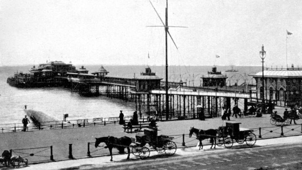 Horse-drawn carriages trot along a seafront with an old Victorian pier visible in the distance. The image is black and white. 