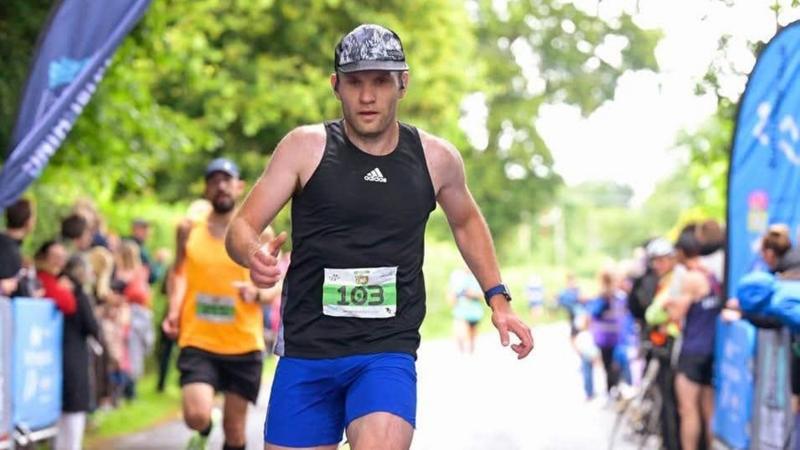 A runner earing a black and white running hat and an Adidas black training top. He is running in a race with a fellow competitor behind him and fans clapping on either side of the track.