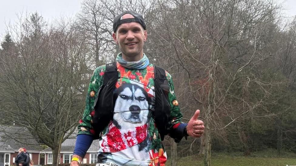 A man has his thumbs up to the camera with a hat facing backwards on his head. he is wearing a Christmas-themed