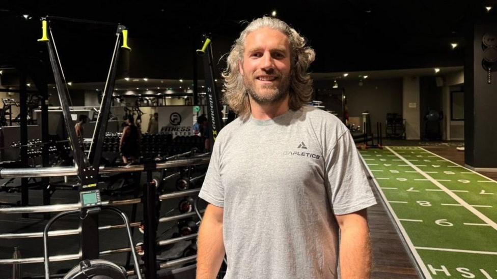 A man stood in a gym wearing a grey t shirt, There are weights on his left and a sprint track on the right.