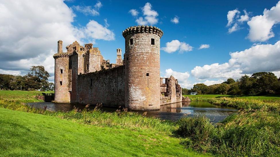 A triangular castle with a moat around it and blue skies with a few clouds above.