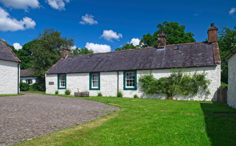 An image of a farmhouse below a blue sky, with green grass and a pavement in front