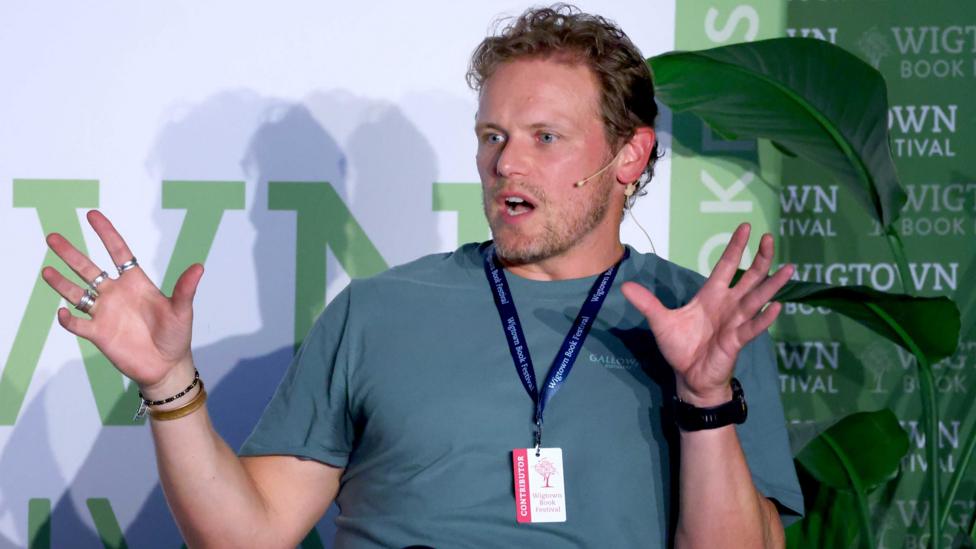 Actor Sam Heughan with a microphone headset on in a bluish green T-shirt in front of a plant at the Wigtown Book Festival. He has a lanyard around his neck.