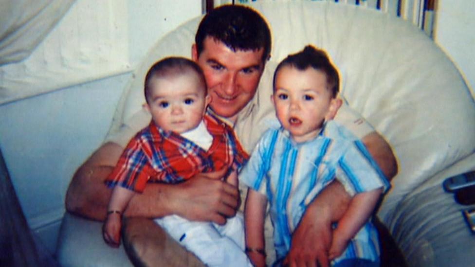 A man with short, dark hair is sitting in a cream leather armchair. He has two young boys sitting on his lap, with an arm wrapped around each. The man, who is looking at the camera, is wearing a cream shirt. The older of the two children is a toddler in a blue shirt. The other is a baby wearing a red flannel shirt. Both boys have dark hair.