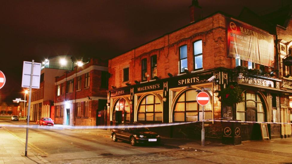 A police cordon stretches across a road between two no entry signs. On the right is the corner view of a bar, which is part of a red-bricked building. The bar has large windows and is painted black with the words 'Magennis's, and 'Spirits' visible. To the left is a well lit street which is largely empty, although a black car is visible near the police tape and a red car is parked further away