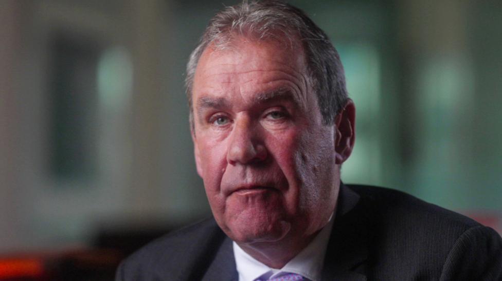 A man with greying dark hair is looking at the camera. He is wearing a dark suit and white shirt with a purple tie just visible at the bottom of the image. He is in a large room with a window, but the background is out of focus.