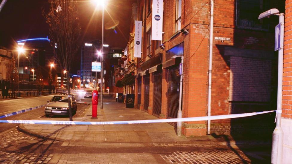 A long street gives way to a cityscape of tall buildings and illuminated street lights. On the right are a series of red brick buildings with an alleyway, which has a sign reading 'market street'. The area outside the alley is cordoned off with police tape which is tied to a lamp post. A silver car and parking meter are seen on the street, which is otherwise empty.