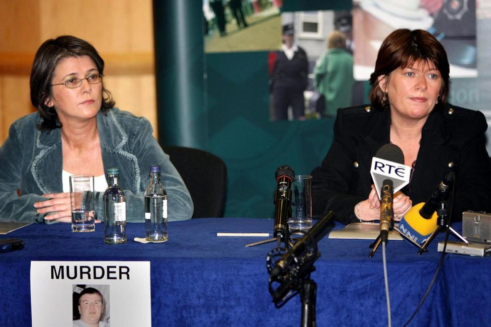 Two women with shoulder-length brown hair sit behind a table at a press conference, with several microphones, glasses of water and bottles placed in front of them. A sign on the front of the table reads “MURDER” above a photograph of a man. Media microphones, including one branded RTÉ, are positioned on the table, while display boards with images are visible in the background.