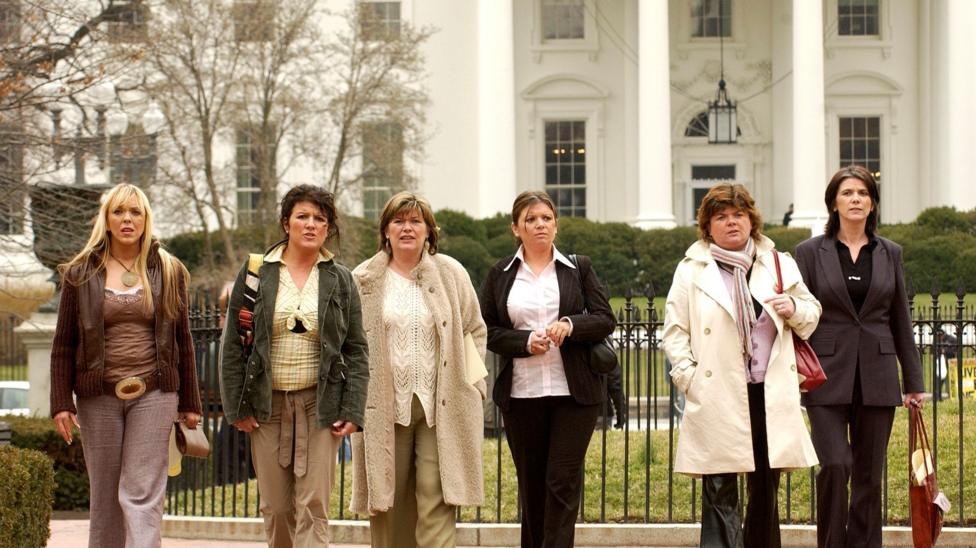 Six women are walking in a broad line towards the camera. Behind them is a white building with large pillars and a black exterior chandelier. There are also neatly trimmed hedges and a garden, surrounded by a black iron fence.